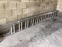 Aluminum extension ladder leaned against a concrete block wall on a rough floor, showing entire ladder length and structure.