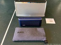 Front view of Bose silver portable speaker and blue AKG Bluetooth speaker with AKG fabric pouch and auction lot tag