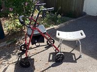 Photo showing red folding mobility walker with a padded seat and black handles, alongside the white shower seat with silver legs on an outdoor pavement.