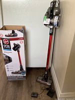 Photo of vacuum cleaner upright with charger and box on floor showing unit, box front, and charging accessories.