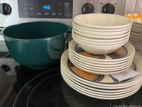 Stack of glazed earthenware dishes with a large green Rosta mixing bowl on a stovetop, showing the variety of plates and bowls stacked.