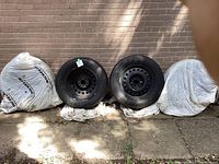 Four tires, two without covers, two covered with white bags, leaning against brick wall outside.