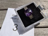 Ring displayed in a clear plastic box with black cushion inside, showing the square cushion cut amethyst colored stone with clear stones on either side.