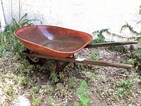 Side view of red metal basin and wooden handles of the wheelbarrow on garden ground.
