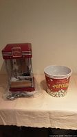 Red Betty Crocker Movie Time popcorn machine on table with one plastic popcorn bucket beside it, machine wrapped partially in plastic.