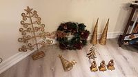Image showing a variety of Christmas decorations placed on a floor including a gold metal spiral tree with star, green wreath with pinecones, two gold cone trees, gold bell, gold decorative tree, and three gold reindeer figurines.