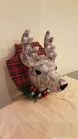 Front view of decorative reindeer head with plaid fabric backing and greenery with berries.