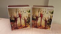 Two unopened boxed sets of glass candle holders, each set containing three pieces with red stems and floral decorations shown on the box.