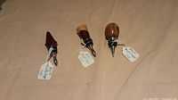 Three handcrafted wooden wine stoppers laid out on a beige surface, showing various wood tops and metal spouts with sealing rings.