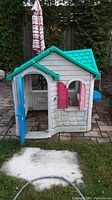 Front and side view of plastic kids outdoor playhouse showing teal roof, cream walls with stone texture, blue door, and red window shutters.