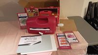 Red Spellbinders Grand Calibur die cutting and embossing machine with handle, manual, cutting plates, and metal dies laid out on pink cloth