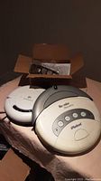 Two iRobot Roomba robotic vacuum cleaners on a table, one older Roomba Discovery showing button controls, the other a newer Roomba with side brush, and a box of accessories behind them.