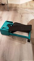 Side angle showing teal green frame and black pedals of the stepper machine on wooden floor.