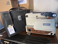 Photo of Majestic Dual Eight 8mm film projector beside its black carrying case, showing logo and wood-tone panel with control dial.
