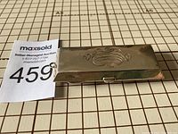 Top-down view showing rectangular sterling silver coin holder with engraved monogram and clasp, placed on grid surface with measurement markings.