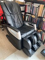 Front view of black massage chair showing overall design, padded seat, backrest, armrests, and foot massager.