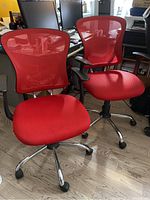 Two red mesh office chairs in an indoor office setting, showing front and side views with surroundings including computer monitors and other office items.