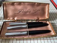 Three vintage Parker 51 pens placed inside satin-lined pen case with the label 'Parker 51' on the interior lid.