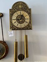 Front view of vintage German wall clock showing ornate brass face with detailed relief decoration, Roman numeral dial, and two cylindrical brass pendulum weights hanging below.