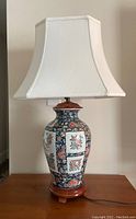 Full view of lamp showing Imari style ceramic body, wooden base, and white pagoda shade, placed on wood surface against a plain background.