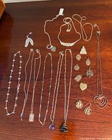 Overview of jewelry on dark wooden surface showing multiple silver color necklaces, feather-shaped earrings with blue bead, various pendants and medals with religious and heart motifs.