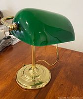 Side view of the bankers lamp showing the green glass shade, brass stem and base, and attached power cord on a wooden surface.