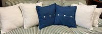 Six throw pillows arranged on a checkered couch. Four white plush pillows and two blue pillows with shell-like buttons.