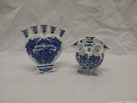 Front view of two Delft tulipieres showing cobalt blue hand-painted designs on white porcelain. Fan-shaped vase on left and round multi-spouted vase on right.