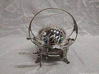 Overall angled view of the silver-plated rotating dish showing the handle, bowl, and one side of the ornate feet. The spreader knife is visible resting on the base, attached by chain.
