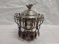 Front view of the antique silver-plated Master Sugar Bowl with 12 spoons hanging around its rim and a bird figurine on the lid.