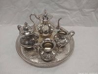 Front view of silver-plated four-piece tea set arranged on round serving tray, showing all pieces with embossed floral details