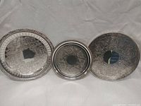 Front view of three silver plated salver trays showing detailed chased and engraved fruit and floral designs on top surfaces with different decorative rims.