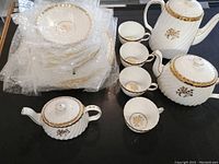 Full view of the tea service showing wrapped dinner and side plates, four teacups, coffee pot, teapot, and creamer with gold floral accents.