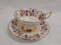 Tea cup and saucer with detailed bramble berry and leaf pattern, gold trim, and scalloped edges, photographed on white background.