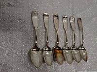 Front view of six Wm Rogers silverware spoons arranged side by side on a foam grey background showing design and condition.