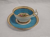 Teacup on saucer showing turquoise blue band with gold trim and interior fruit motif design