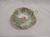 Single teacup on saucer showing detailed gold leaf pattern and pink rose center