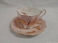 Tea cup on saucer with detailed view of pink base, gold and brown wheat botanical pattern, and gilded rims.