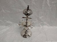 Front view of vintage silver-plated spoon holder with bell showing base, spoon hooks, central column and bell with black knob.