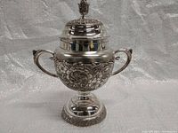 Front angled view of the ornate silver-plated sugar bowl showing embossed floral details, two handles, and lid finial.