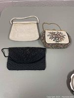 Three vintage clutches displayed on a grey surface: a white beaded Walborg clutch, a black beaded La Royale clutch, and a floral tapestry La Marquise clutch.