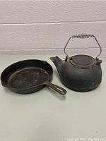 Photo showing cast iron frying pan and wood stove kettle on white surface against brick wall.
