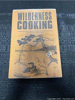 Front cover of the Wilderness Cooking hardcover cookbook from 1973 with illustrated campfire scene and title text.