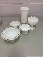Five white milk glass pieces including three open loop rim bowls, tall fluted vase, and hobnail textured bowl on pedestal bases, arranged on gray table in front of pale pink wall.