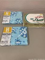 Three vintage sheets in original packaging on a tabletop: two blue floral Heritage double flat sheets and one white twin flat sheet in beige and white packaging.