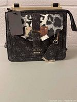 Front view of new Guess handbag with visible tags, black and grey logo pattern, animal print panel, and gold-tone clasp.