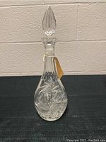 Full view of clear crystal wine decanter with stopper and tag, placed against a tiled wall on a black surface.