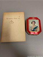 Photo showing the front cover of the book and the collectors tin with Queen Elizabeth II portrait side by side on a grey surface.