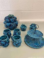 Full view of all items in the lot including four mugs, a two-tiered serving tray, a lidded container, a small pitcher, and a fruit-shaped decorative piece, all in blue ceramic with fruit relief designs.