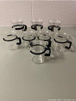Front angle of 5 clear glass coffee mugs with black handles and rims displayed on a white surface.